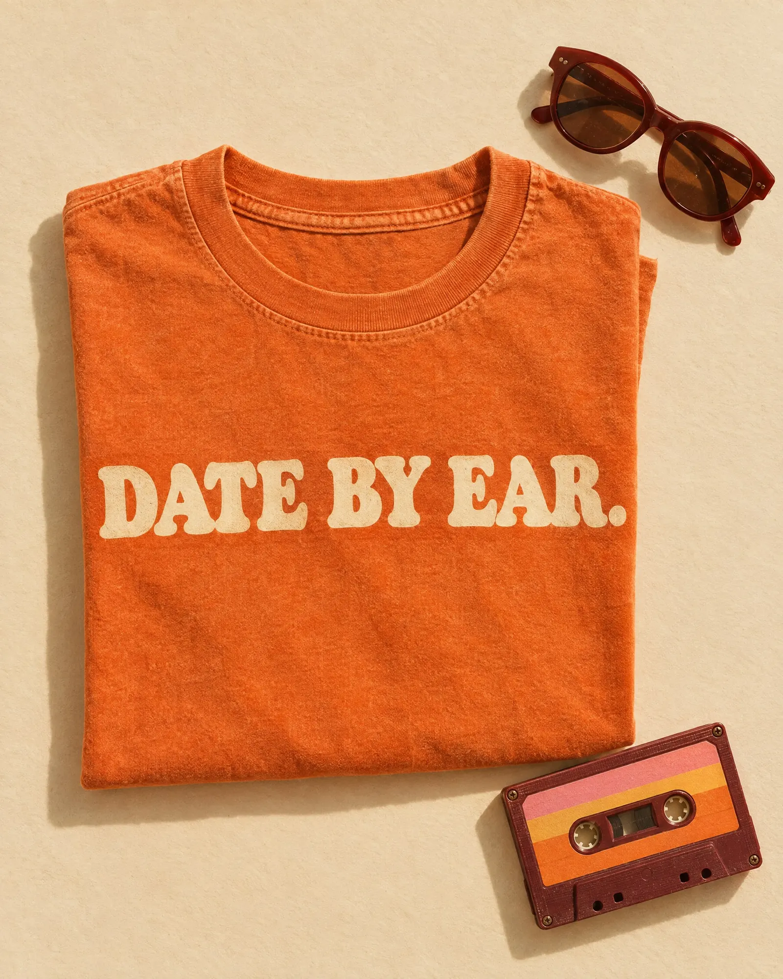 A folded tangerine T-shirt printed “Date by ear.” next to sunglasses and a sunset-striped cassette.