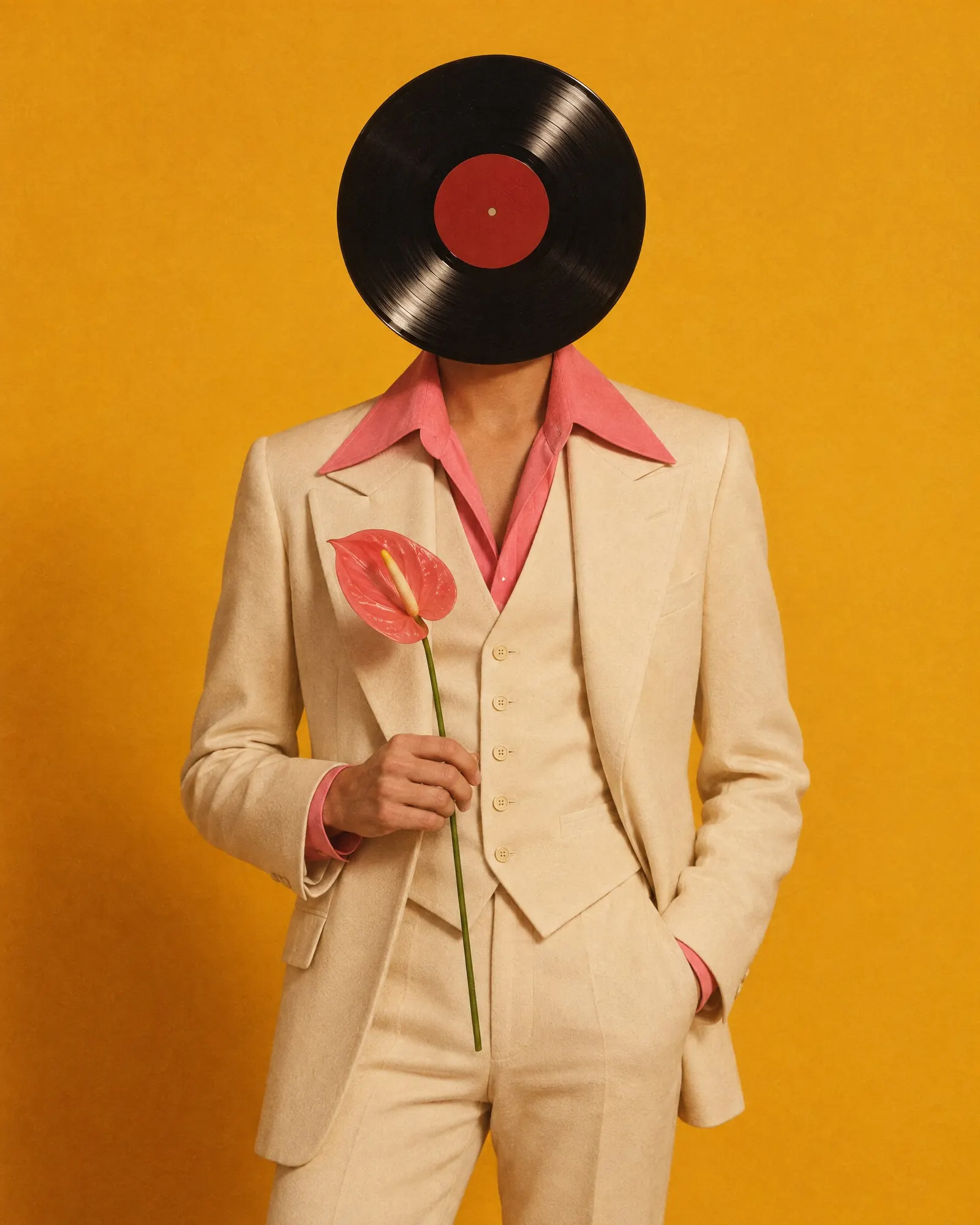 A Record Head in a cream three-piece suit with a pink collar holds a single pink flower against a marigold backdrop.