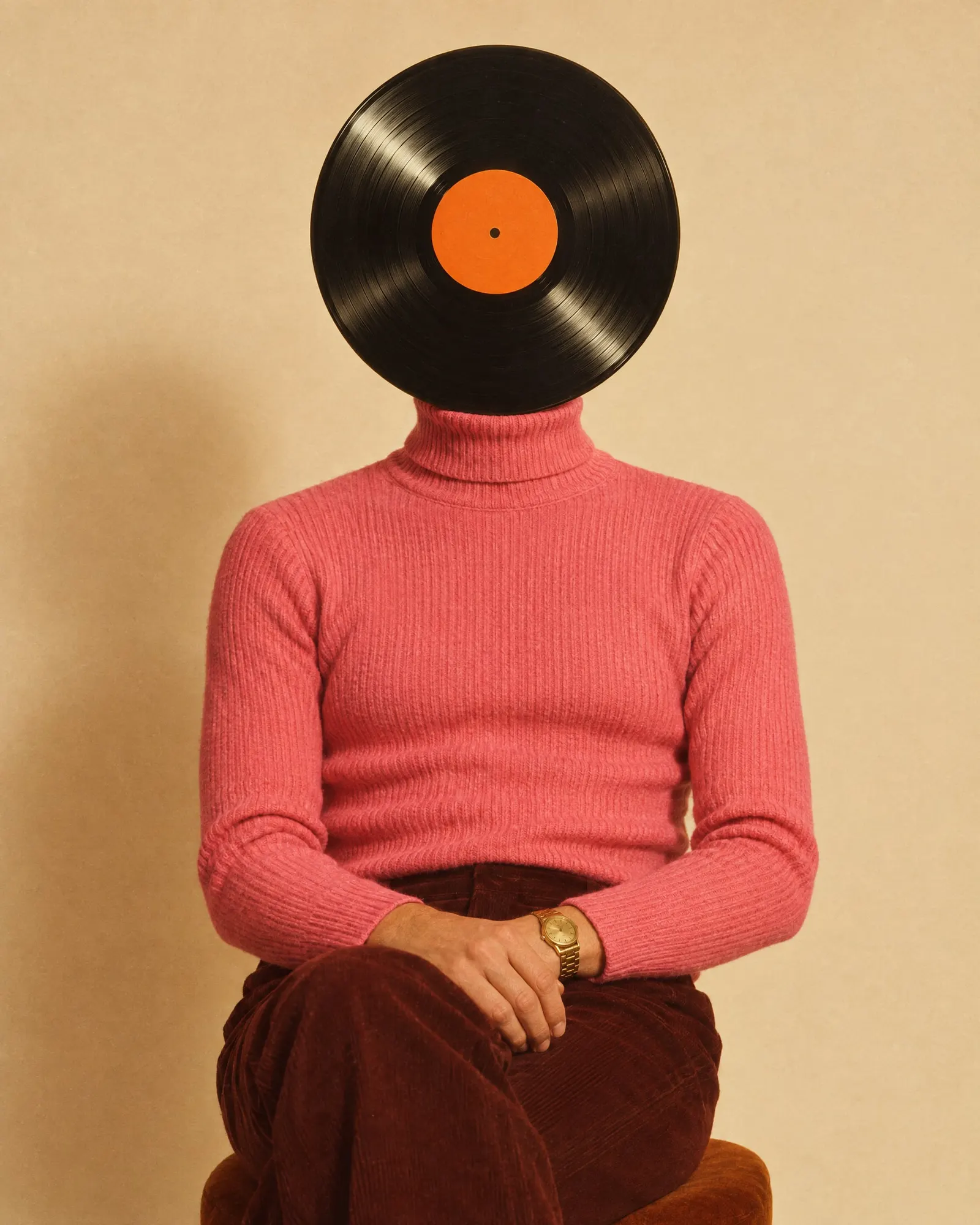 A Record Head in a pink ribbed turtleneck and a gold watch sits against a cream backdrop, an orange-labelled record where the face would be.